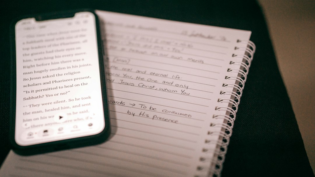 A smartphone displaying religious text beside a spiral notebook with handwritten notes